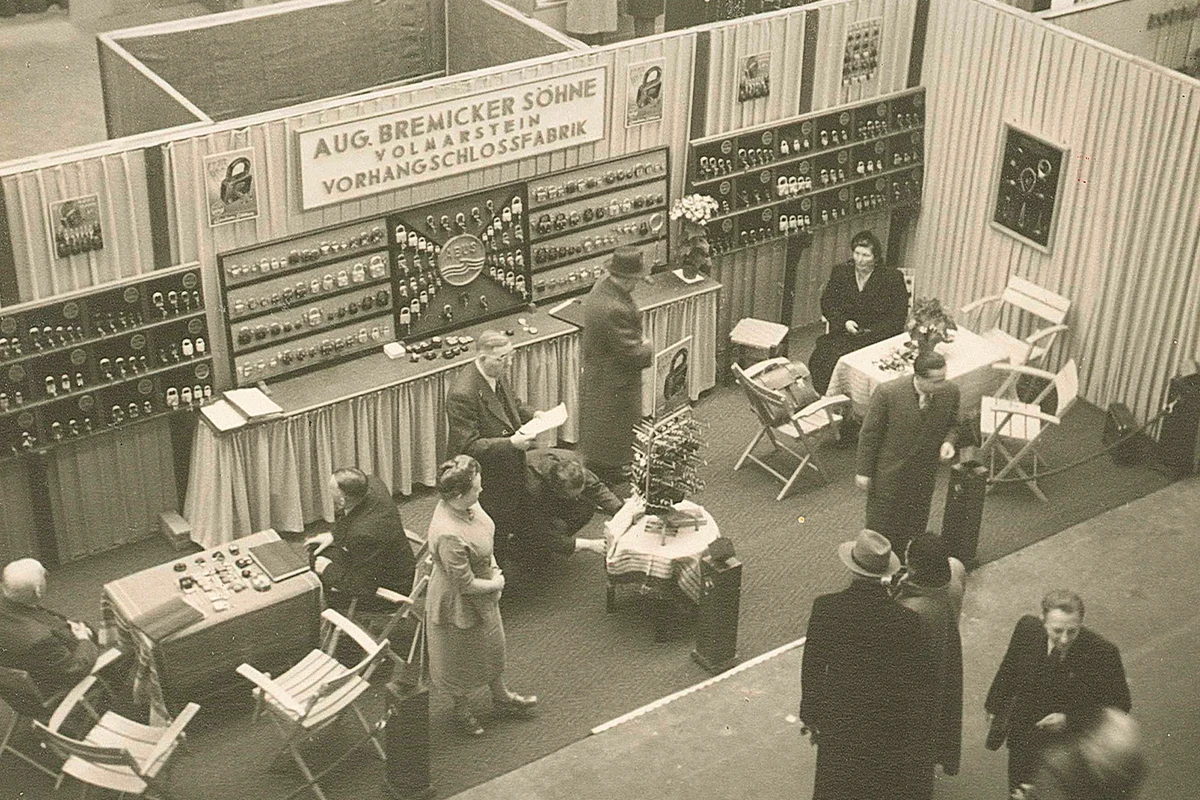 Ein Messestand in den 1950er Jahren, mit Vorhangschlössern, die an den Wänden hängend gezeigt werden und einigen Tischen, an denen Menschen sitzen oder stehen, die sich teilweise unterhalten © ABUS