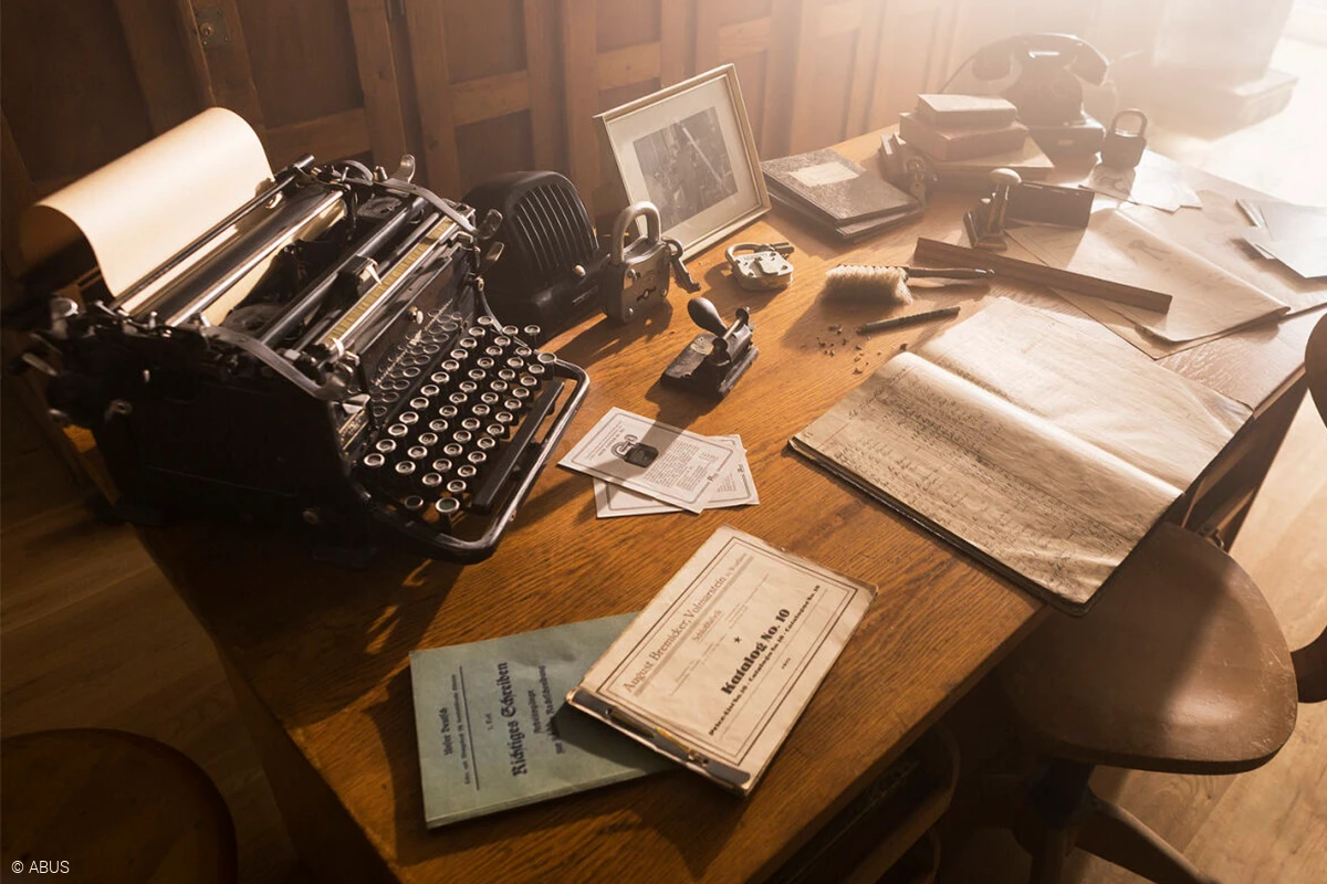 Ett skrivbord i trä med olika mappar och dokument, en skrivmaskin, en gammal telefon, en tavelram och enstaka gamla hänglås © ABUS