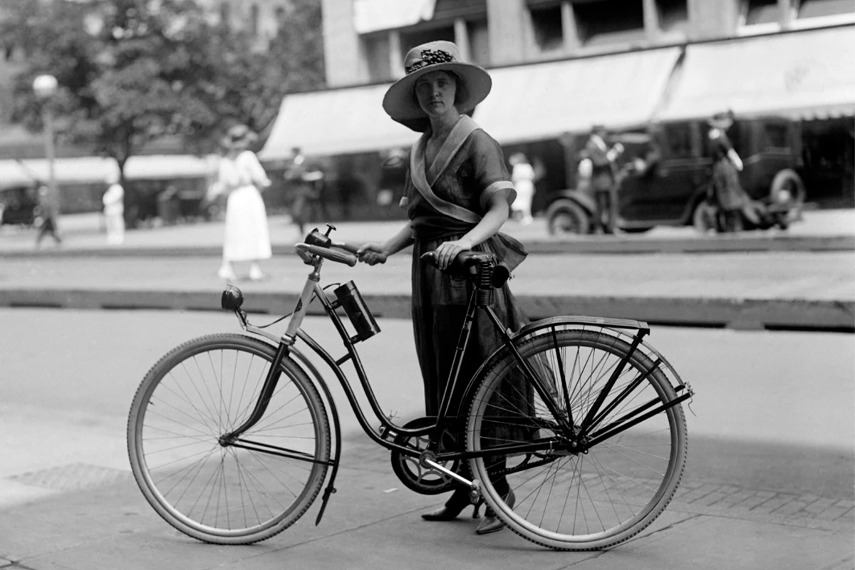 A lady in a hat stands behind a ladies' bicycle and looks directly into the camera. Her surroundings are blurred © shutterstock - Everett Collection