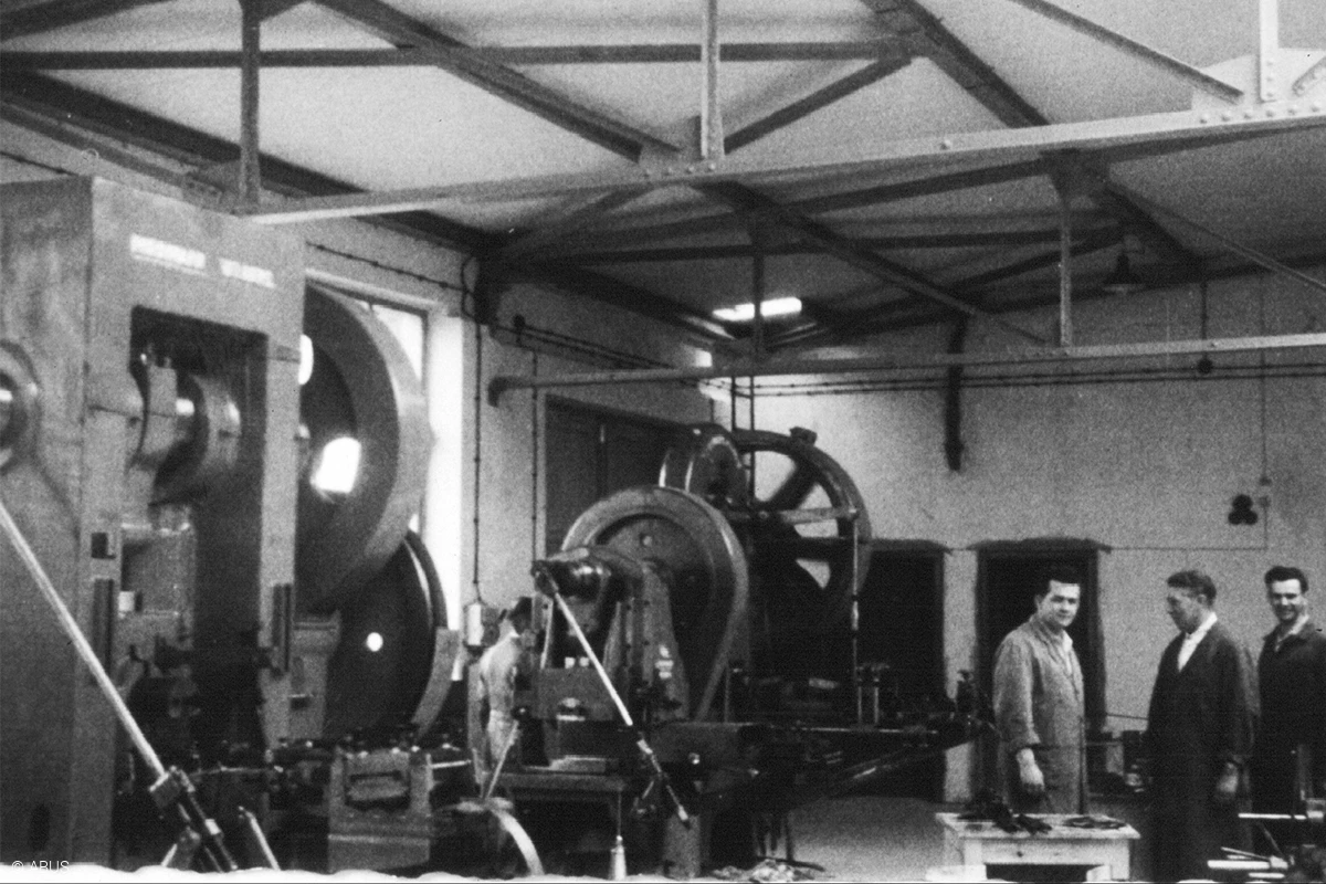 En del av en produktionshall som visar några av maskinerna till vänster och tre män som samtalar till höger © ABUS
